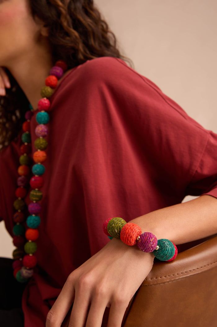 Person wearing a red shirt, colorful cotton-wrapped wooden beads necklace, and the Rainbow Kantha Spheres Bracelet, resting their arm on a brown chair.