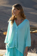 A woman stands outdoors in a sunlit, desert-like setting, wearing the Relaxed Aqua Cardigan with a boxy silhouette and a colorful beaded necklace.