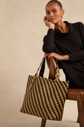 A woman in a black dress sits on a chair, resting her elbow on the backrest, holding the Reversible Braided Shopper—a spacious handbag featuring a striking black and tan checker pattern.