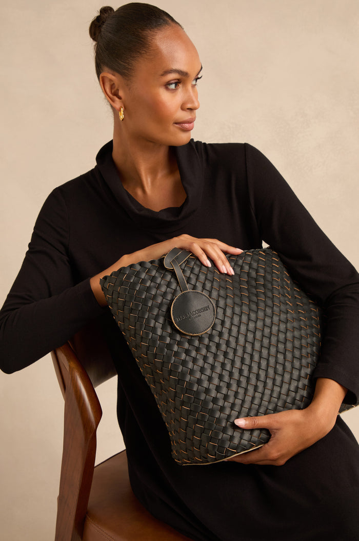 Woman in a black dress sits on a wooden chair, holding a large woven reversible bag with a round tag.