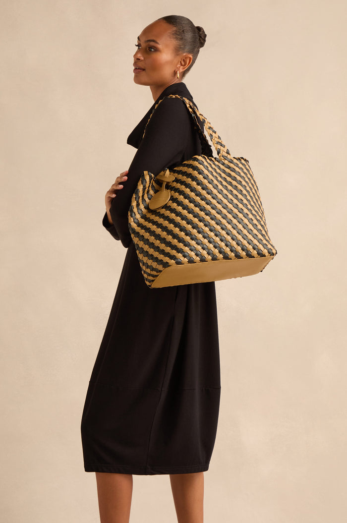 A woman in a black dress stands sideways, carrying the Reversible Braided Shopper in bold black and tan hues over her shoulder against a neutral background.