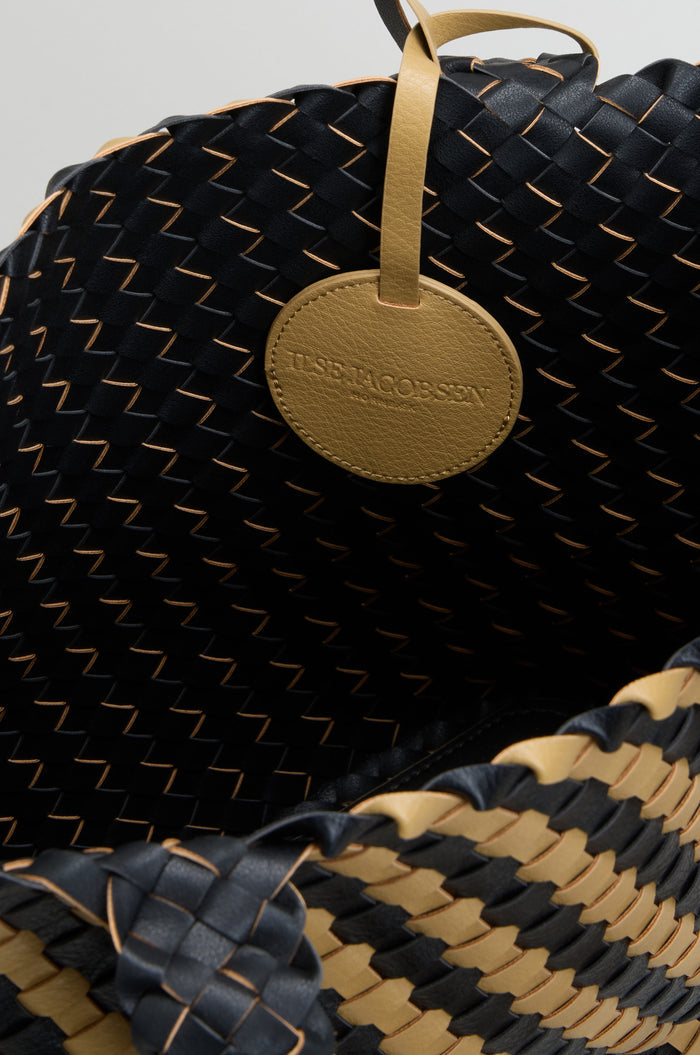 Close-up of a basket weave handbag in black and tan, featuring a round tan leather tag embossed with “ILSE JACOBSEN.”.