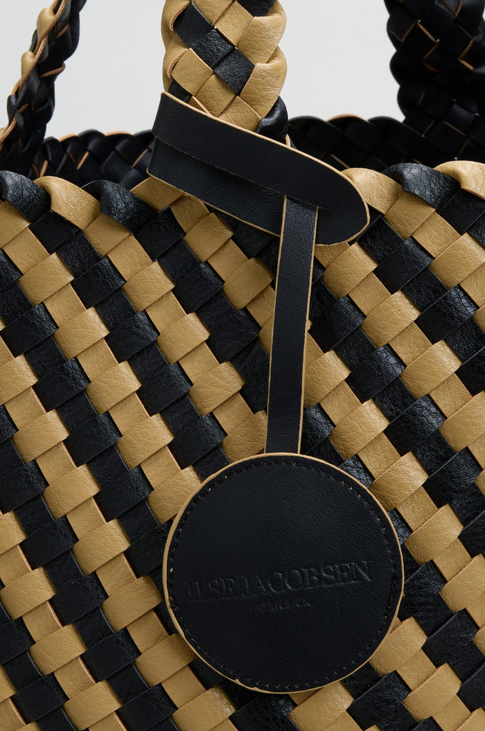 Close-up of the Reversible Braided Shopper, featuring a woven black and tan basket weave design and a round black leather tag labeled “ILSE JACOBSEN HORNBÆK.”.