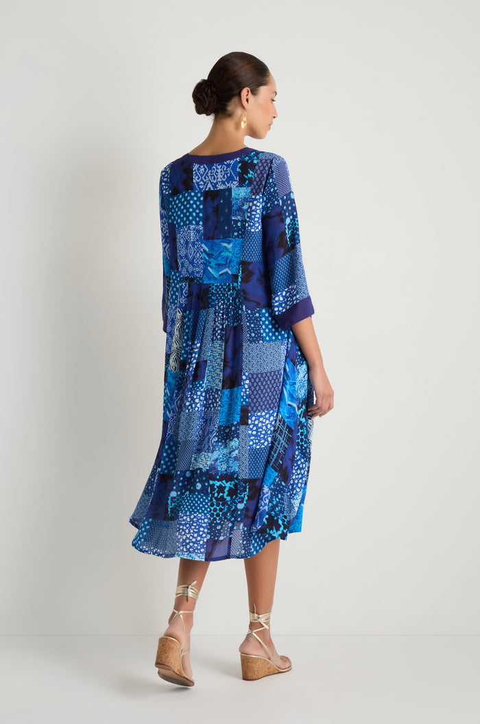 A woman stands against a plain background, facing away, wearing the Sahara Indigo Patchwork Flared Dress in viscose crepe with a flared, mid-length silhouette and half sleeves, paired with wedge sandals.