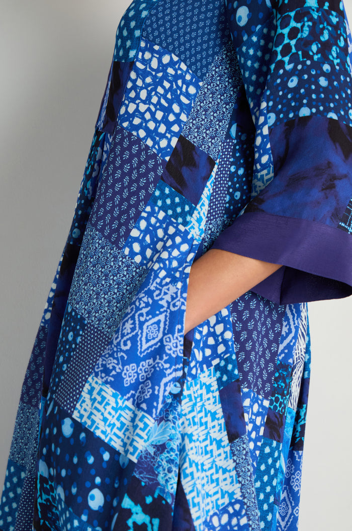 Someone wears the Sahara Indigo Patchwork Flared Dress, made from viscose crepe with blue geometric and abstract patchwork patterns, their hand tucked into a side pocket.