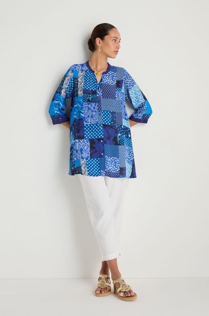 A woman stands against a white wall, wearing the Sahara Indigo Patchwork Longline Shirt in soft viscose crepe, paired with white pants and beige sandals, arms relaxed and head turned to the side.