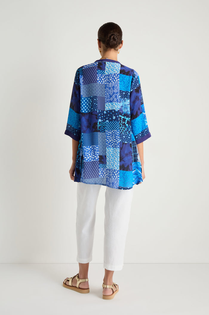 A person faces away, wearing the Sahara Indigo Patchwork Longline Shirt with white pants and beige sandals, set against a plain white background.