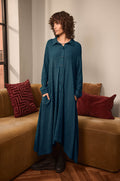 A person stands indoors on a tan sofa, wearing the Seersucker Squares Shirt Dress—a long, teal checkered design made in England—paired with black boots and hands tucked into pockets. Decorative pillows accent the sofa, and a window is visible in the background.