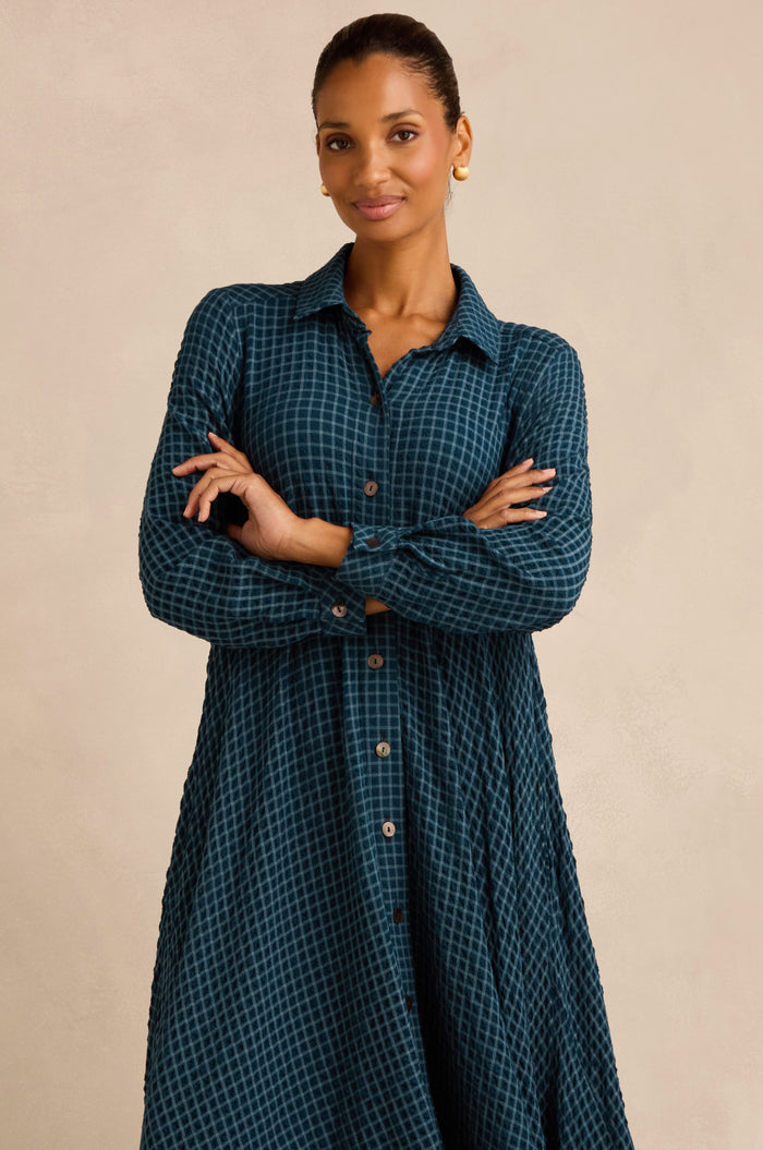 A woman stands with her arms crossed, smiling slightly as she models the Seersucker Squares Shirt Dress in a teal and black check pattern, paired with gold hoop earrings, set against a plain beige background. This chic dress is crafted in England.