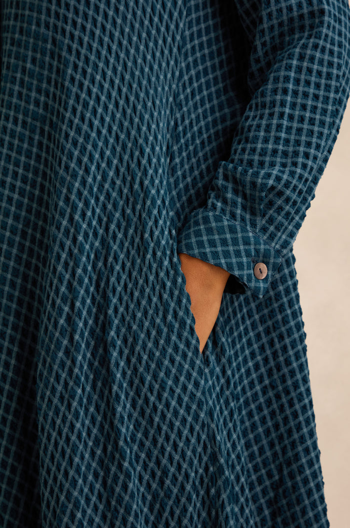 Close-up of a person wearing the Seersucker Squares Shirt Dress, featuring textured blue checkered fabric, with one hand in the side pocket and the sleeve cuff secured with a button.
