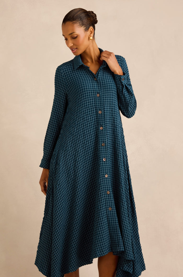 A woman wearing the Seersucker Squares Shirt Dress—a teal plaid, long-sleeved dress made in England—stands against a plain beige background, looking downward.