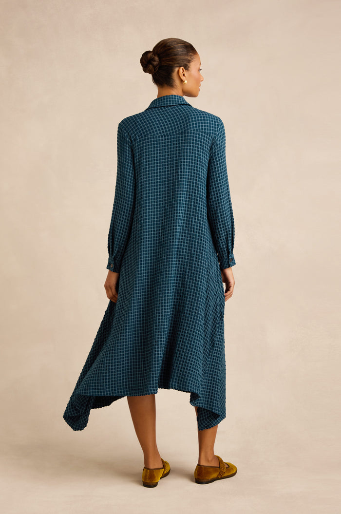 A woman stands with her back to the camera, wearing the Seersucker Squares Shirt Dress—featuring long sleeves, a blue checked pattern, and an uneven hem—paired with yellow shoes against a neutral background.