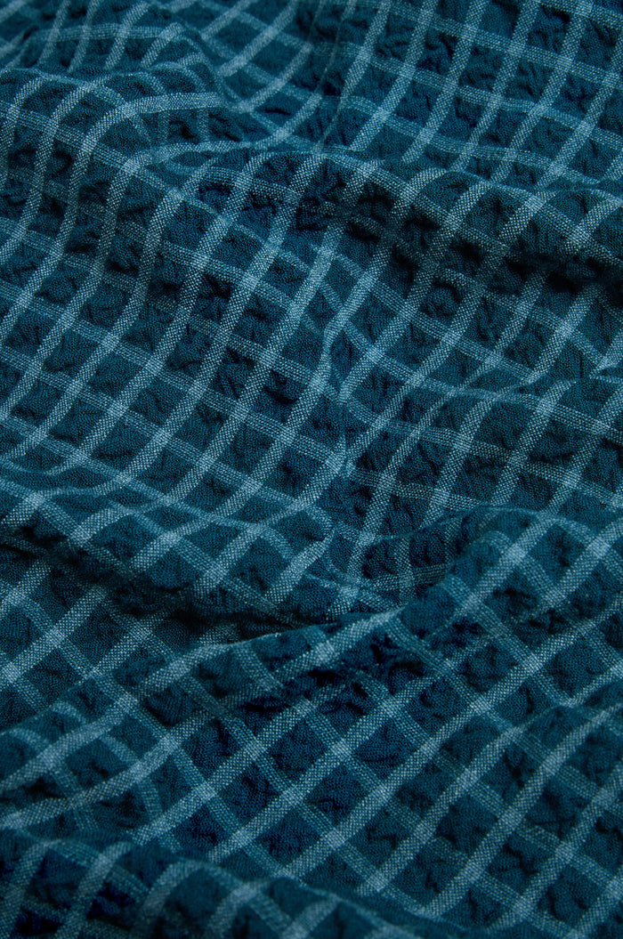 Close-up of the Seersucker Squares Shirt Dress, featuring blue fabric with a textured crisscross grid pattern reminiscent of classic seersucker. The material looks soft and gently puckered—ideal for this shirt dress made in England.