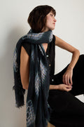 A woman in a sleeveless black outfit sits on a stool, wearing the Shibori Wool Scarf draped over her shoulders. The breathable, patterned dark scarf contrasts elegantly as she looks to the side against a plain white background.