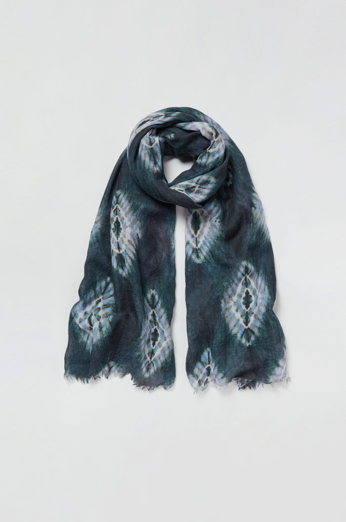 The Shibori Wool Scarf features a dark, tie-dye inspired pattern and fringed edges, creating a breathable and stylish accessory when laid flat on a white background.