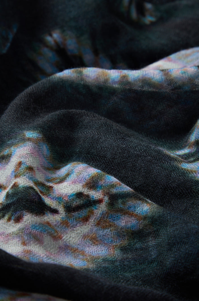 A close-up of the Shibori Wool Scarf reveals a textured fabric with a multicolored abstract pattern, featuring dark, wavy folds and subtle hints of blue, pink, and white. This scarf showcases a breathable design that blends artisanal style with comfort.