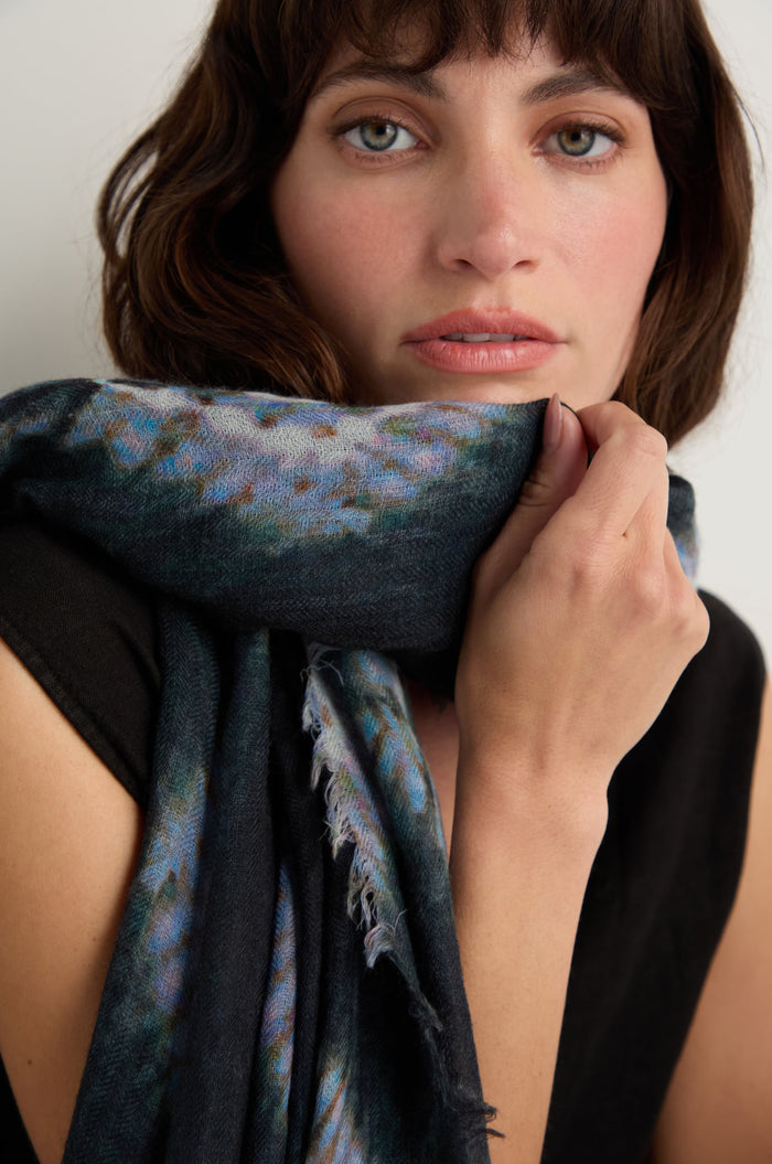 A woman with wavy brown hair holds the Shibori Wool Scarf close to her face. Wearing a black top, she gazes directly at the camera against a plain background, highlighting the breathable elegance of this accessory.
