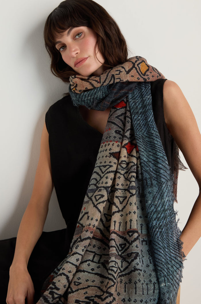 A woman in a sleeveless black top wears the Sketch Mountain Wool Scarf—a large, lightweight wool accessory featuring geometric and abstract print designs in muted colors—while sitting against a light background.