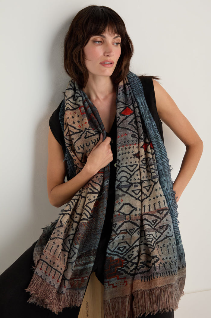 A woman wearing a sleeveless black top and the Sketch Mountain Wool Scarf—a large, lightweight wool scarf with fringe and an abstract print—stands against a plain white wall.