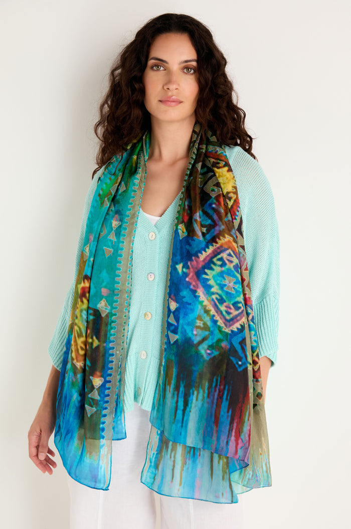 A woman models the Stained Glass Silk Scarf with a turquoise sweater and white pants, standing against a simple light-colored background.