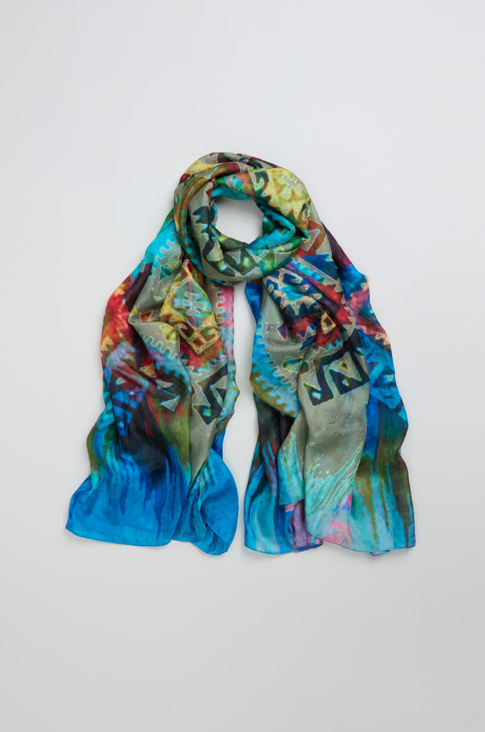 The Stained Glass Silk Scarf features colorful abstract patterns in blue, green, red, and yellow tones, arranged in a loose loop on a plain light background.