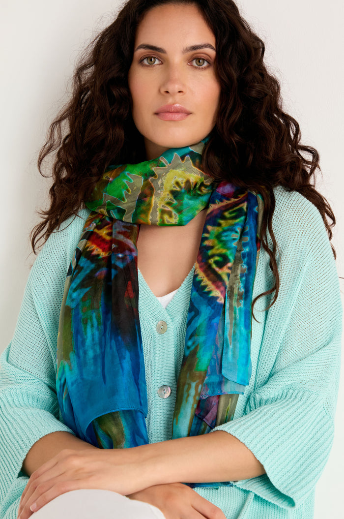 Woman with long curly hair wears a light blue sweater and the Stained Glass Silk Scarf, featuring an abstract print, while sitting against a plain light background.