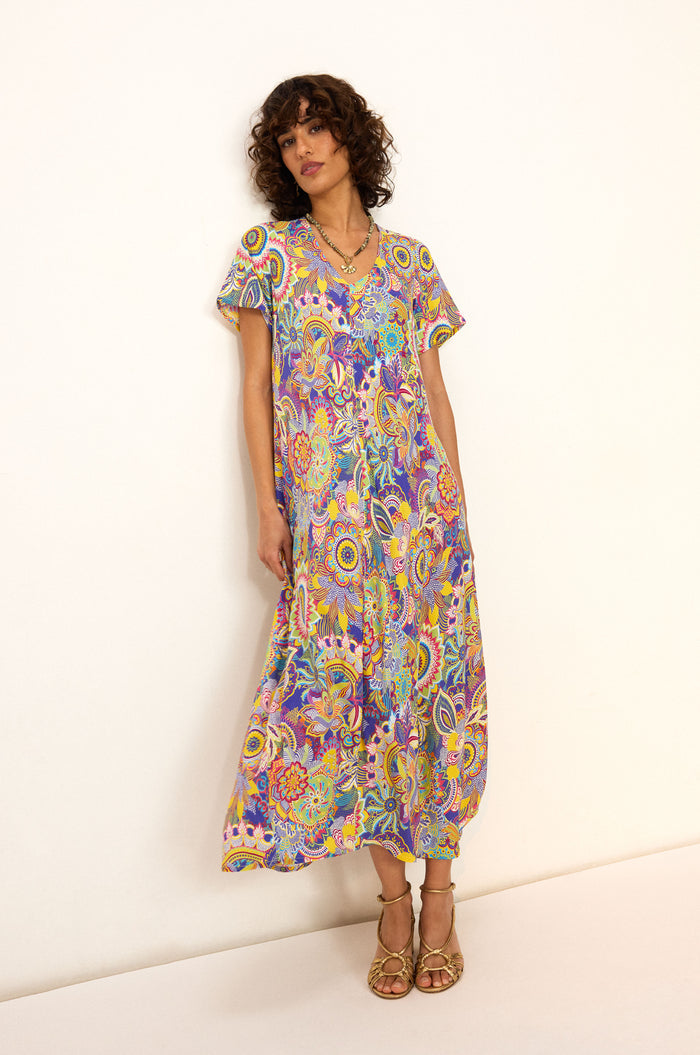 A woman stands against a white wall wearing the Summer Sarasa Bubble Dress—an ankle-length, short-sleeve dress with a colorful paisley print in silk-like viscose—paired with tan sandals.