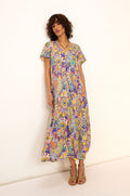 A woman stands against a white wall wearing the Summer Sarasa Bubble Dress—an ankle-length, short-sleeve dress with a colorful paisley print in silk-like viscose—paired with tan sandals.