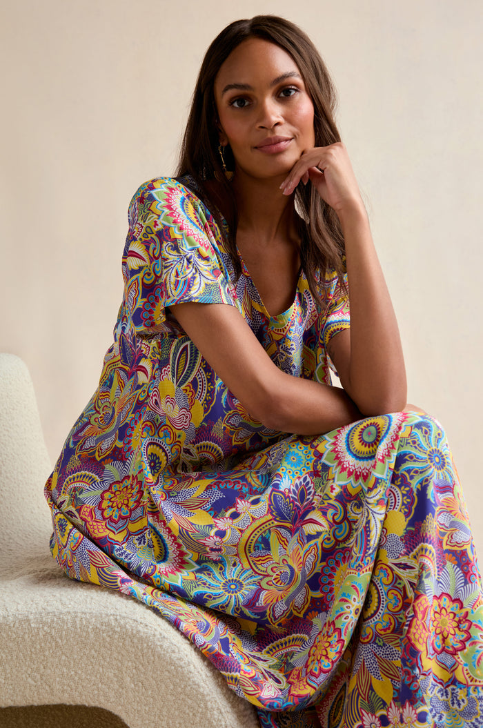 A woman sits on a chair, wearing the Summer Sarasa Bubble Dress, featuring a vibrant floral and paisley print in silk-like viscose, and gazes at the camera with a relaxed expression.