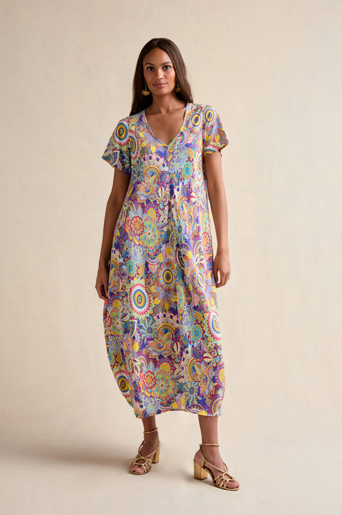 A woman stands against a neutral background wearing the Summer Sarasa Bubble Dress, a loose-fitting, colorful floral-print piece with short sleeves made from silk-like viscose, paired with gold sandals.