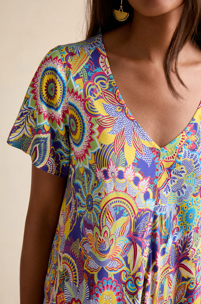 A woman wears the Summer Sarasa Bubble Dress, a colorful V-neck with short sleeves, featuring bold floral, paisley, and geometric patterns in silk-like viscose.