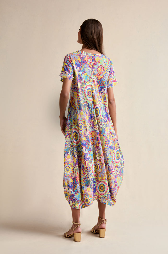 A woman faces away, wearing the Summer Sarasa Bubble Dress with a vibrant paisley print and beige heeled sandals against a plain background.