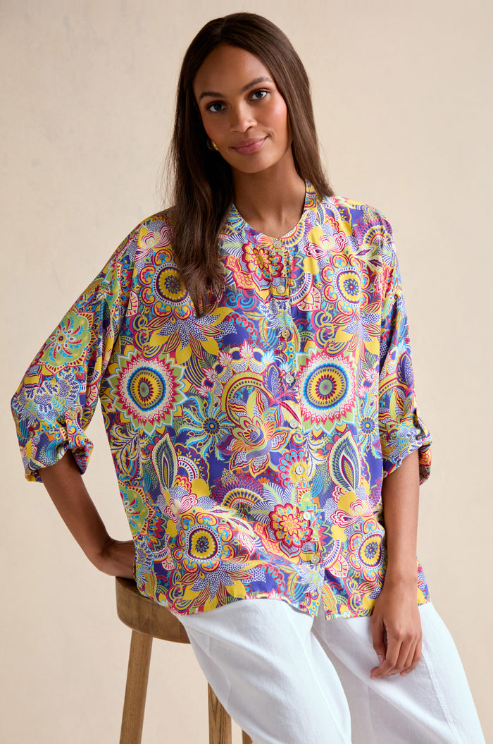 A woman with long brown hair wears the Summer Sarasa Relaxed Shirt paired with white pants as she sits on a wooden stool against a plain beige background.