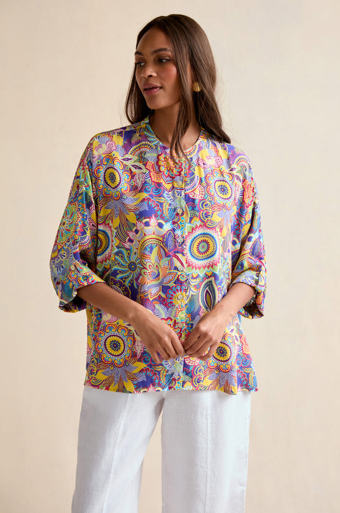 A woman wears the Summer Sarasa Relaxed Shirt, an oversized, colorful blouse with a mandala pattern, paired with white pants and posed against a simple, light background.