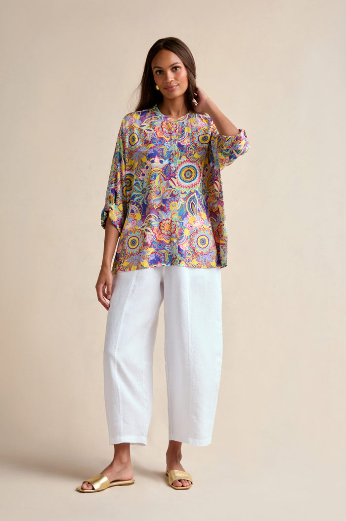 A woman stands against a plain background wearing the Summer Sarasa Relaxed Shirt, styled with loose white pants and gold flat sandals.
