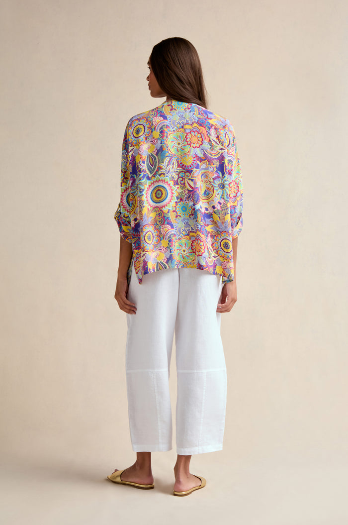 A woman stands facing away, wearing the Summer Sarasa Relaxed Shirt with a boxy silhouette, white wide-leg pants, and beige flat shoes against a plain beige background.