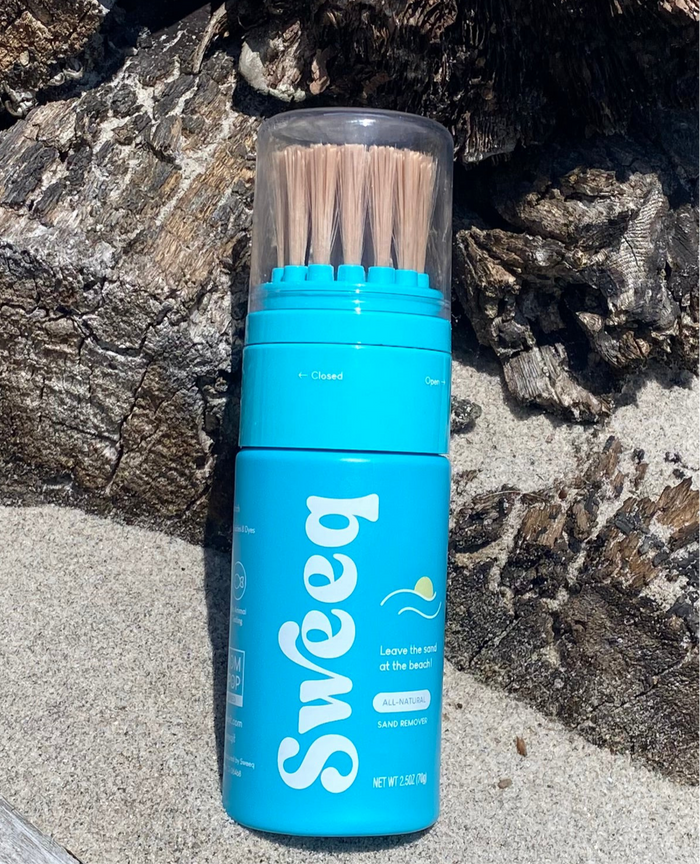 Front view of Sweeq Beach Sand Remover showcasing wooden bristles and blue bottle on a rocky beach surface