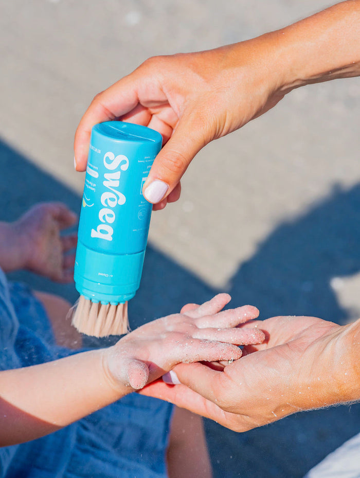 Person applying sunscreen to a child's hand using a blue sand remover stick with a brush