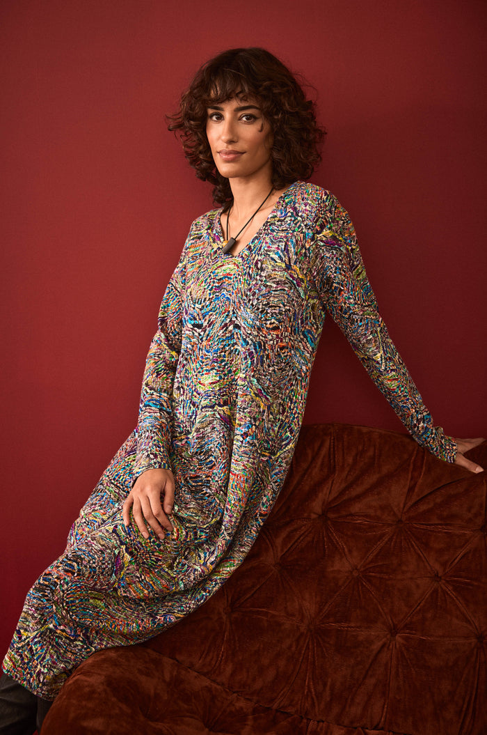 A person with curly hair wearing the Textured Prism Jersey Bubble Dress sits on a brown cushioned couch against a dark red wall.