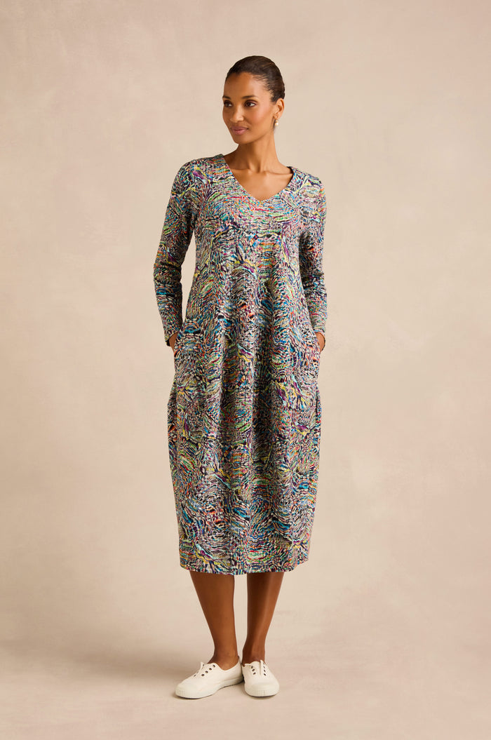 A woman stands against a neutral background wearing the colorful Patchwork Fields Jersey Bubble Dress, featuring long sleeves, convenient pockets, and white sneakers.