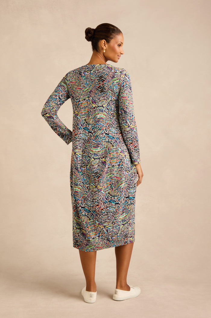 A woman stands with her back to the camera, wearing a patterned Patchwork Fields Jersey Bubble Dress with long sleeves and white slip-on shoes against a neutral background.