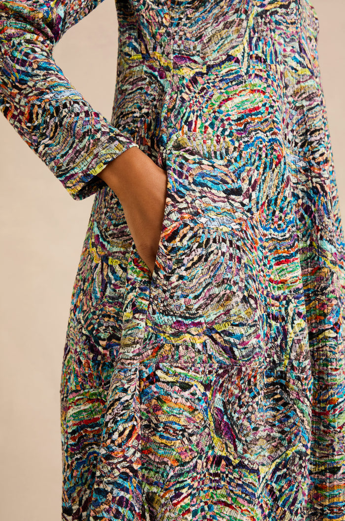 A person with medium skin tone wears a vibrant Patchwork Fields Jersey Bubble Dress with long sleeves and a colorful abstract pattern, casually resting one hand in the dress pocket.