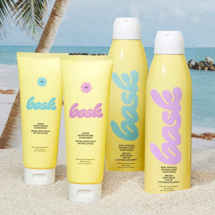 Four pastel yellow and blue skincare bottles labeled 'bask' in script, arranged on a white surface with a palm leaf.