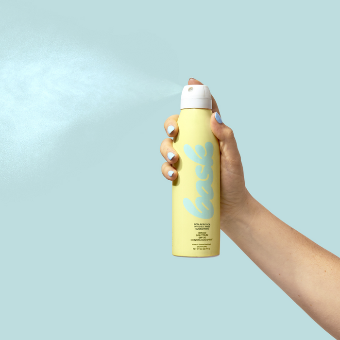 Hand holding a yellow sunscreen bask spray can against a pale blue background