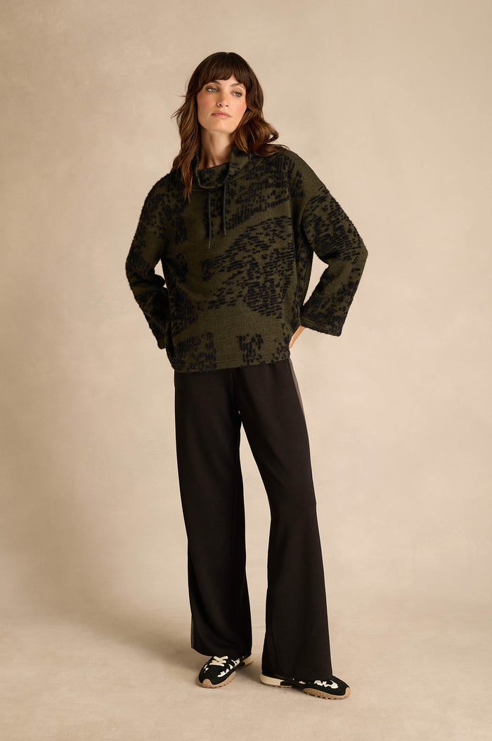 A woman stands against a neutral background, wearing the Thyme Speckled Jersey Boxy Top in green and black with a striking pattern, paired with black wide-leg pants and black slip-on shoes featuring white accents.