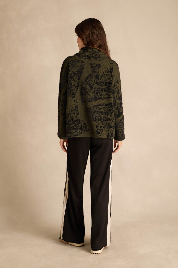 A person stands with their back to the camera, wearing the Thyme Speckled Jersey Boxy Top—a dark green, patterned jacket-style top—paired with black pants featuring white side stripes, set against a neutral background.
