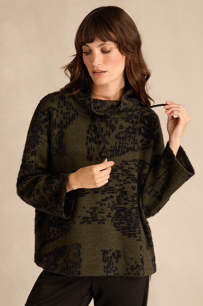 Woman wearing the Thyme Speckled Jersey Boxy Top, featuring a textured dark green and black pattern, boxy fit, and wide sleeves, looking down while holding one side of the collar.
