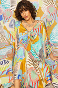A woman with curly hair sits on a patterned surface, wearing a Tropical Fantasy Kaftan Style Dress with an abstract tropical print and necklace, blending seamlessly into the matching background.