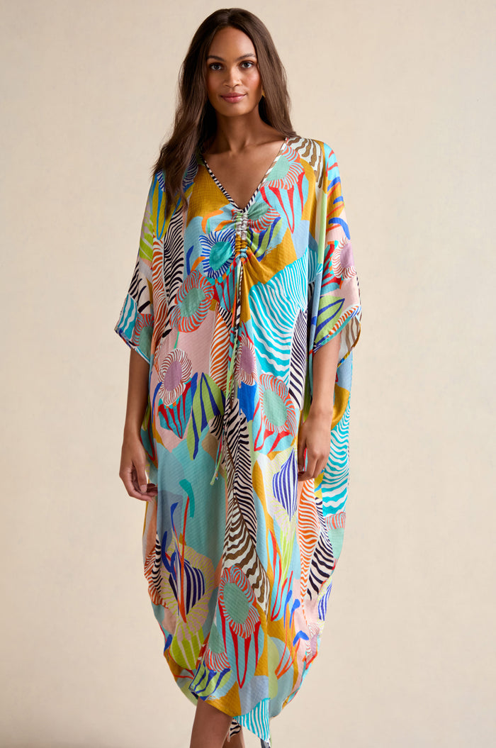 A woman wears a loose, colorful Tropical Fantasy Kaftan featuring a V-neck and short sleeves, standing against a plain background.