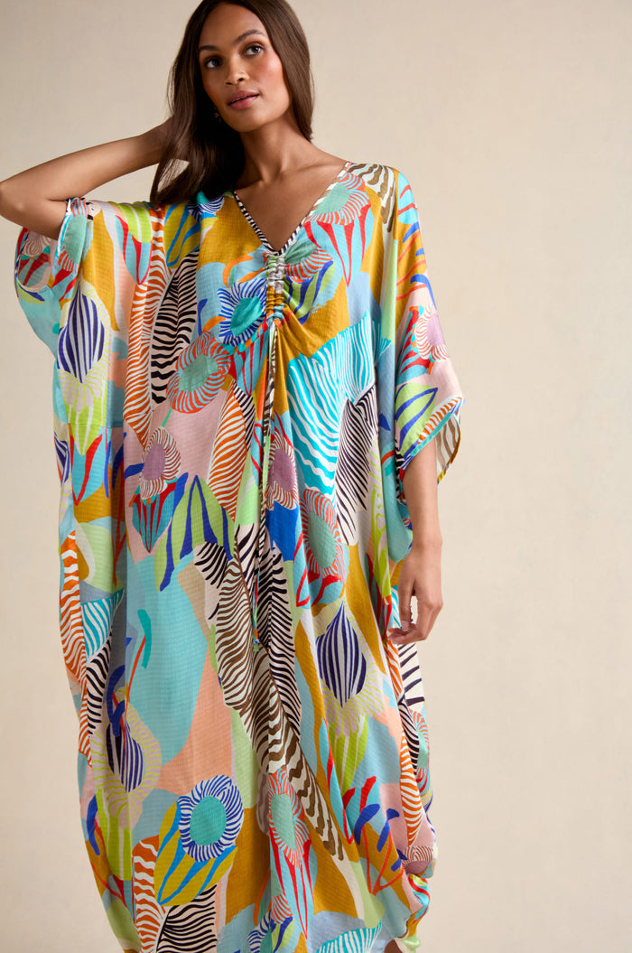 Woman wearing the Tropical Fantasy Kaftan, an oversized maxi dress with abstract, multicolored patterns, poses with one hand touching her hair against a plain background.